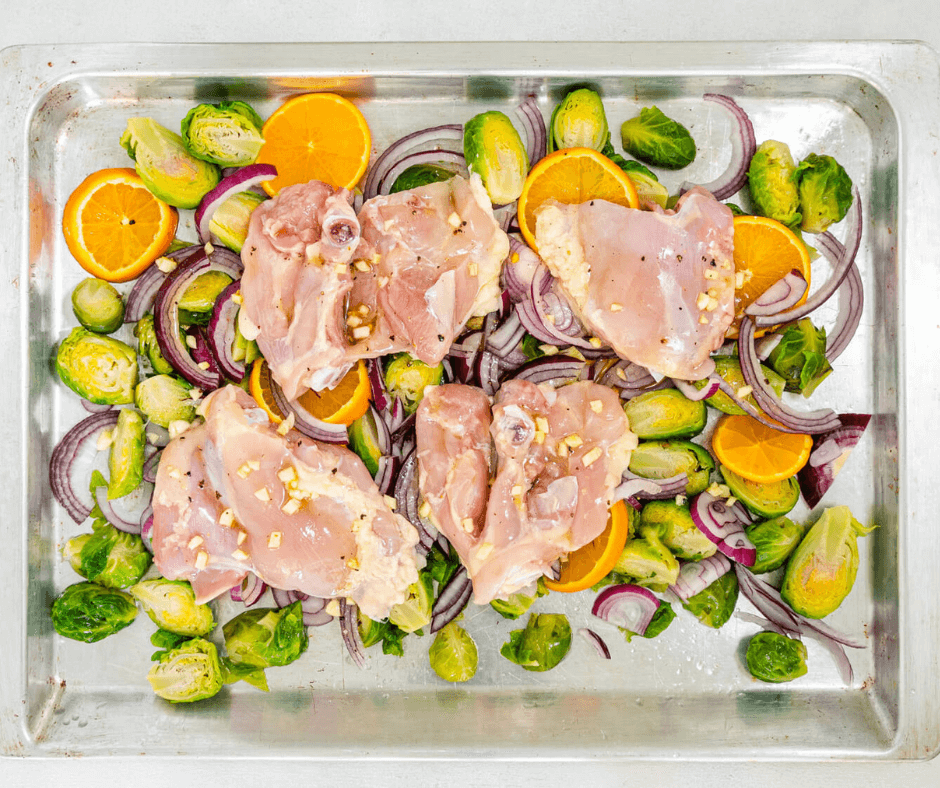 Brussel sprouts, orange slices, and red onion around chicken thighs on a baking sheet